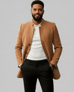 Man wearing a brown coat over a white shirt and dark pants on a gray background