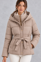 Women’s belted hooded quilted puffer jacket with warm padding, a high neckline, adjustable tie waist and practical side pockets.