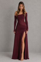 Woman wearing a burgundy off-shoulder gown with a high slit on a gray background