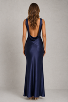 A woman viewed from the back wearing a navy blue satin maxi dress with a deep open back and cowl neckline, standing against a light gray studio background.