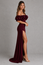 Woman wearing a burgundy off-shoulder gown with ruffles and a high slit on a gray background
