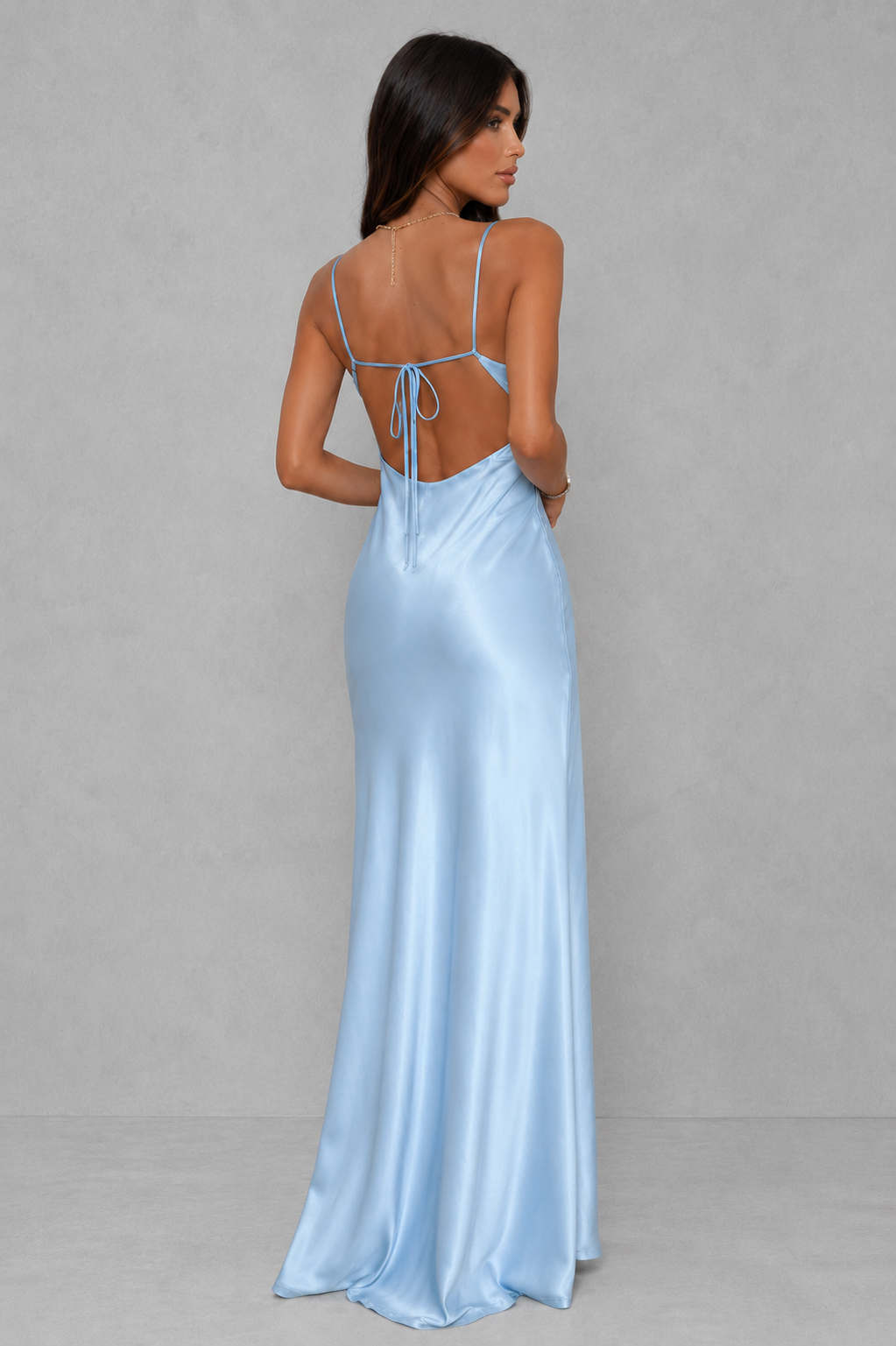 A woman modeling a light blue satin maxi dress from the back, featuring thin spaghetti straps, a tie-back neckline, and a floor-length flowing skirt, set against a neutral gray studio background.