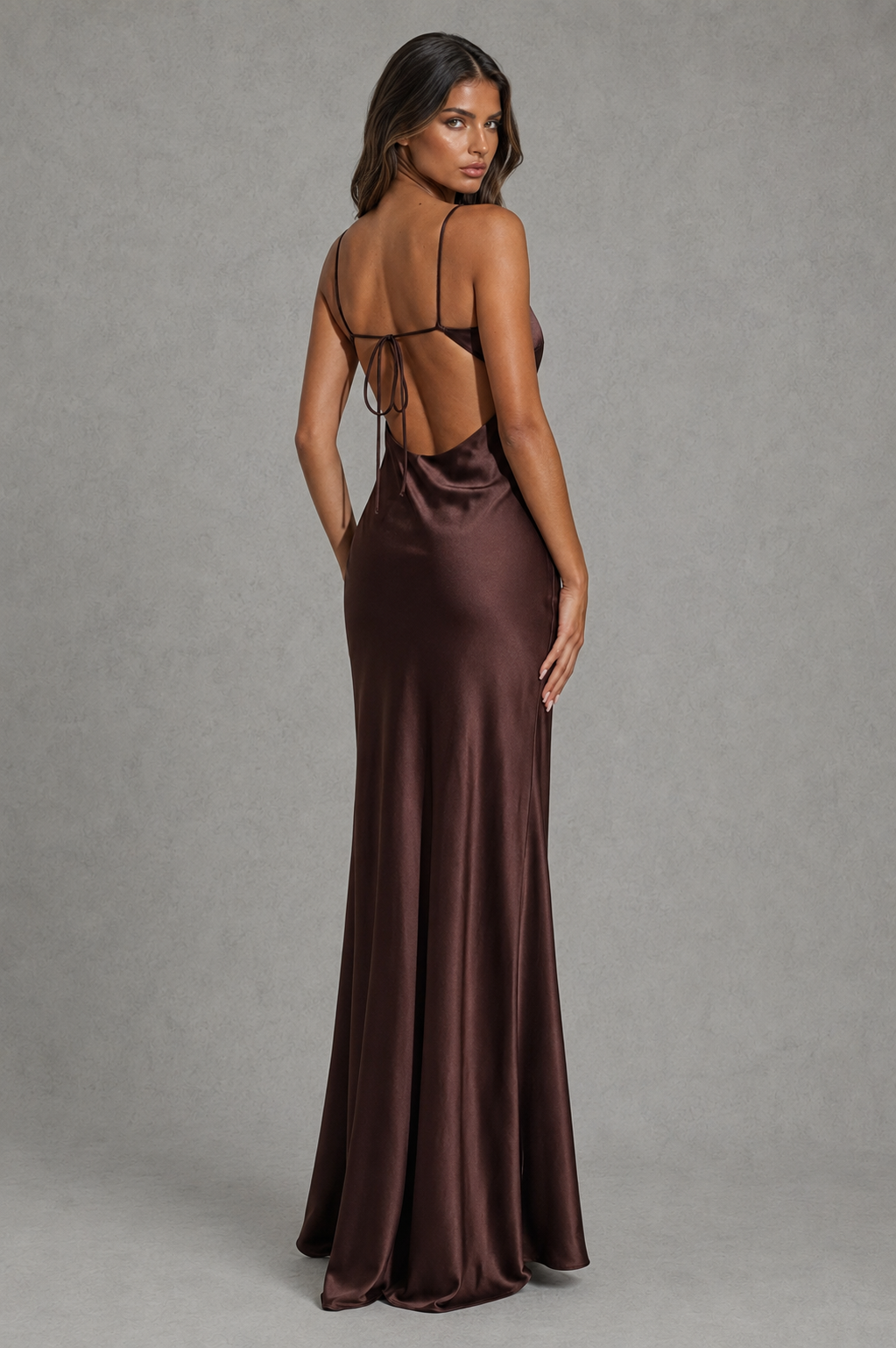 A woman modeling a floor-length satin gown from the back, showing a deep open back with thin tie straps. The dress is a rich dark brown color with a satin sheen, set against a neutral gray studio background.
