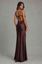 A woman modeling a floor-length satin gown from the back, showing a deep open back with thin tie straps. The dress is a rich dark brown color with a satin sheen, set against a neutral gray studio background.