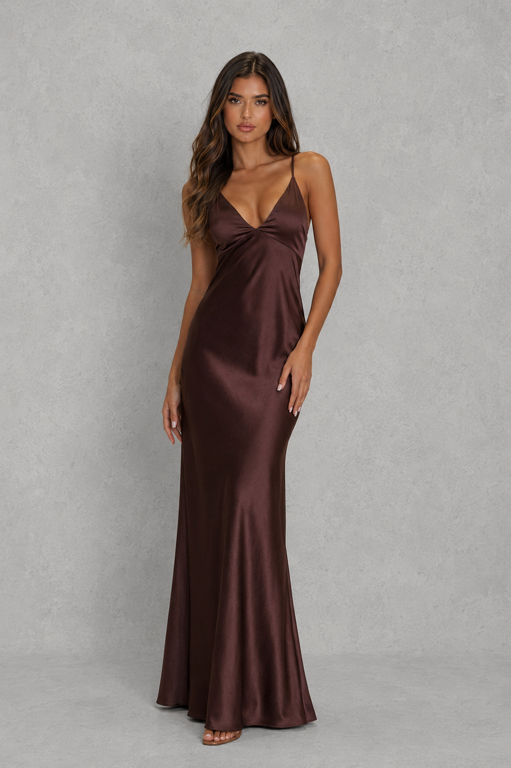 A woman modeling a floor-length satin slip dress in a deep chocolate brown color, standing against a textured gray studio backdrop.