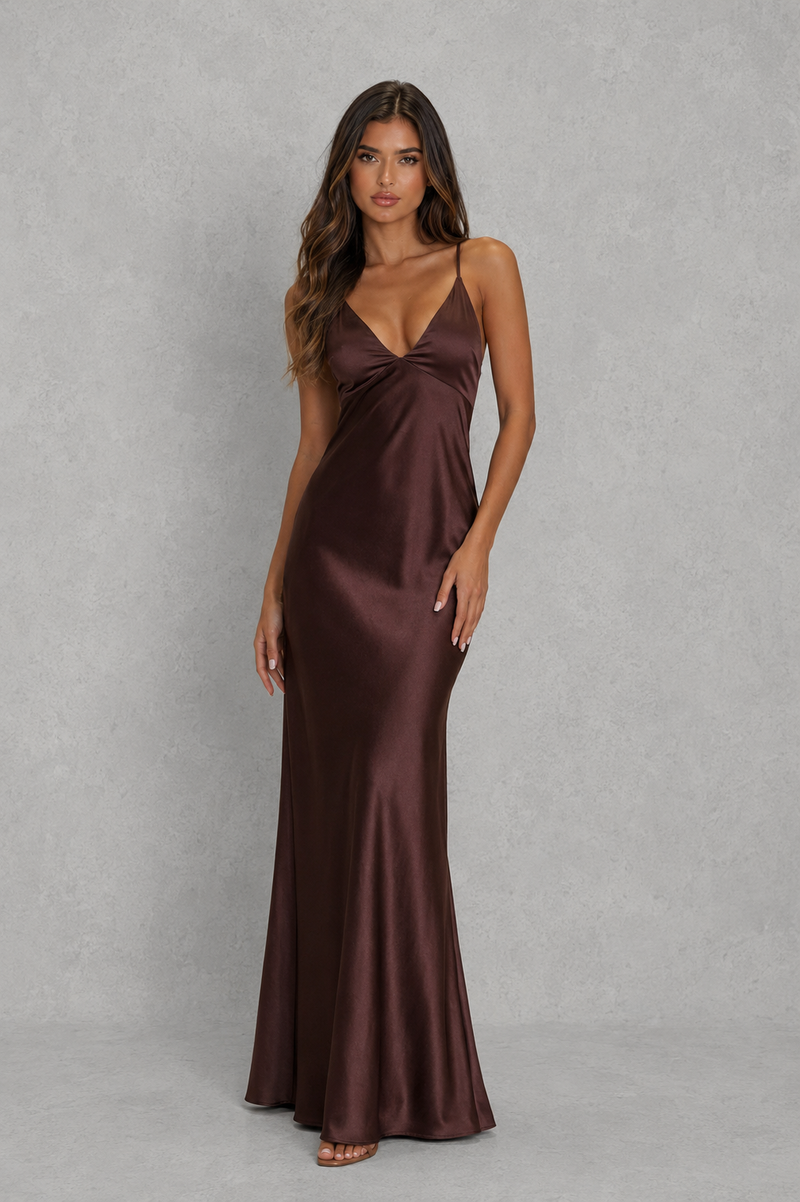 A woman modeling a floor-length satin slip dress in a deep chocolate brown color, standing against a textured gray studio backdrop.