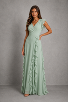 A woman modeling a sage green maxi dress with ruffle details, standing against a textured gray studio backdrop.