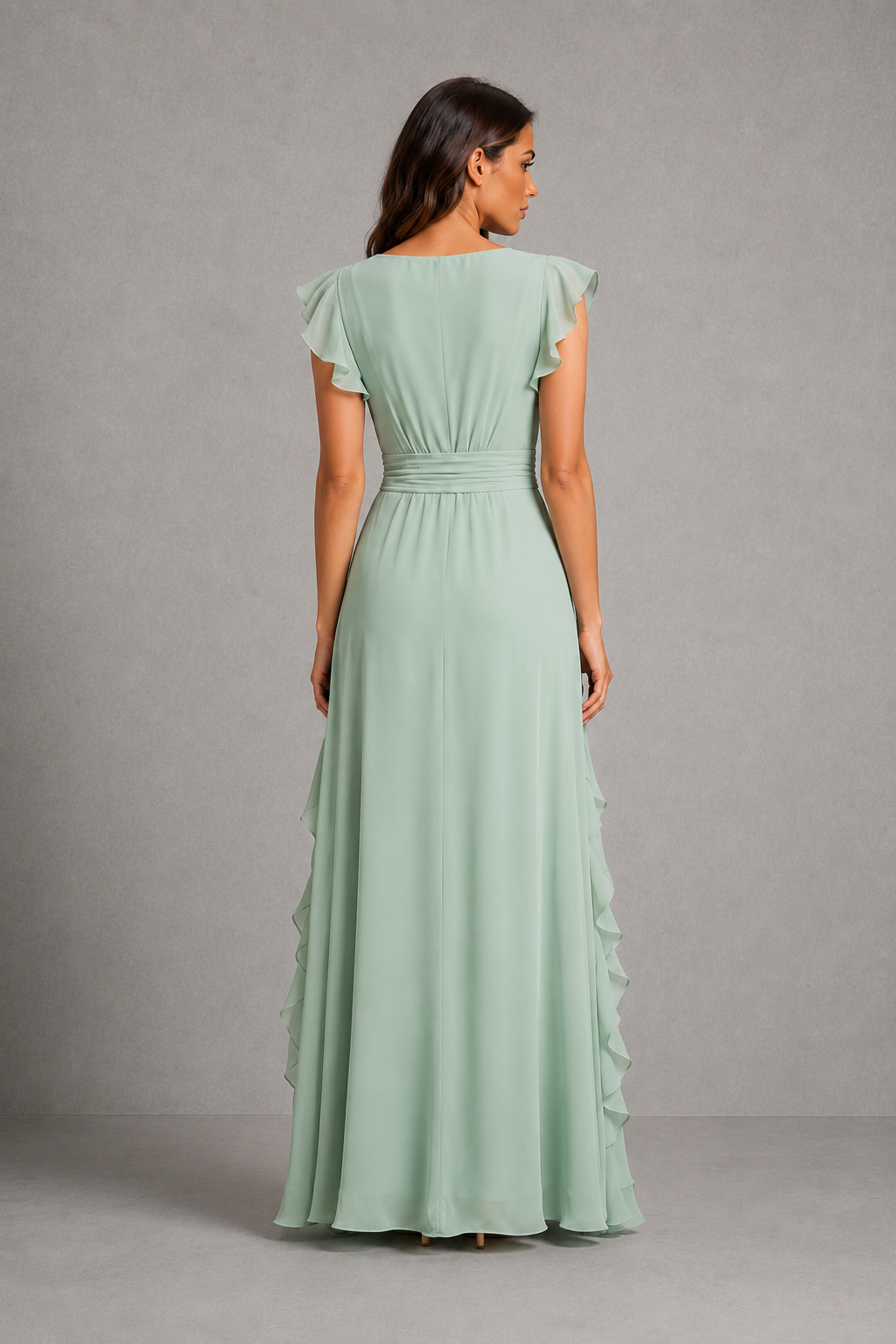 A woman viewed from the back wearing a floor-length sage green dress with ruffled cap sleeves, a gathered waist, and layered ruffle detailing along the skirt, photographed against a plain gray studio backdrop.