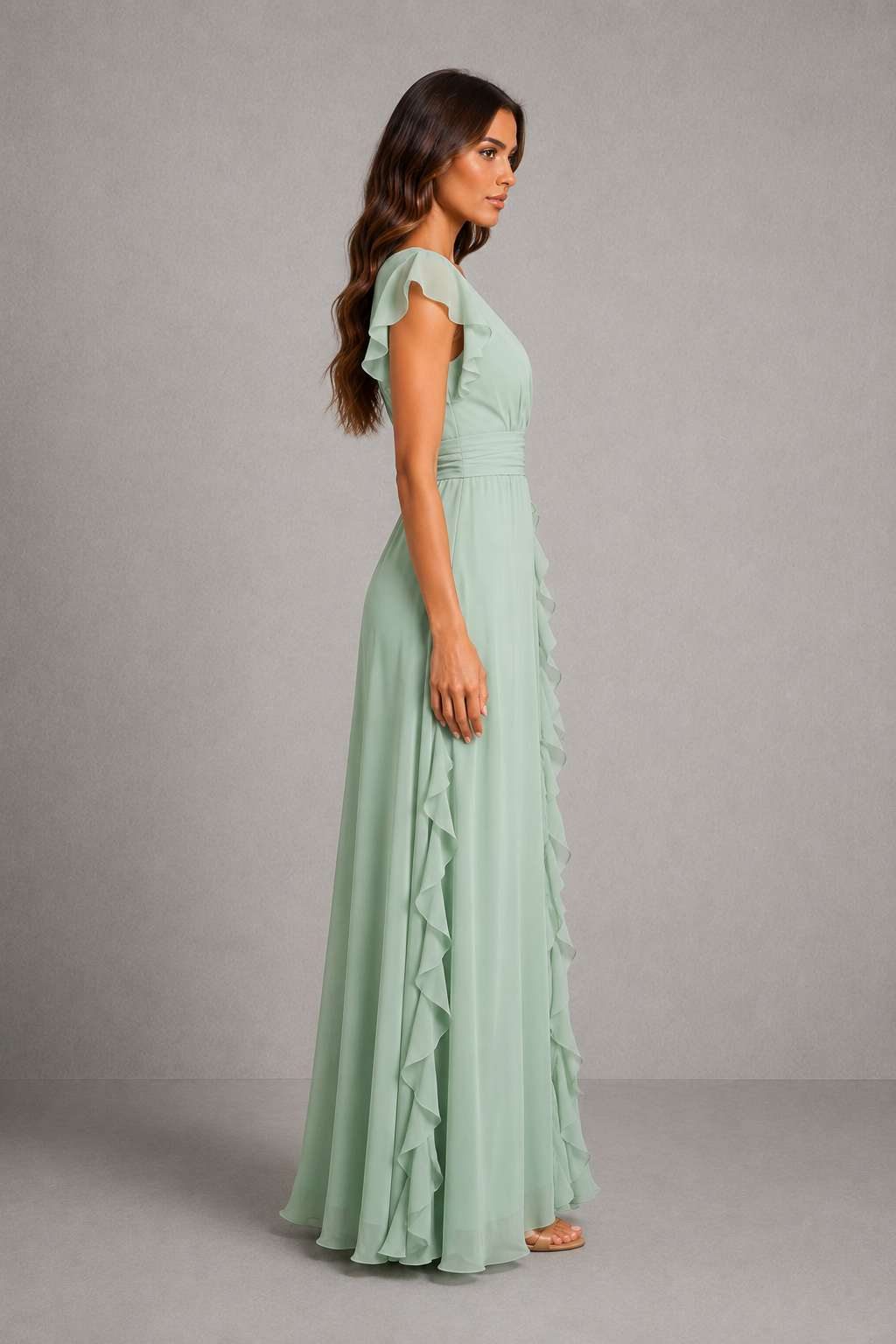 A woman modeling a floor-length sage green dress with ruffle details, shown from a side profile against a neutral gray studio background.