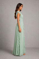 A woman modeling a floor-length sage green dress with ruffle details, shown from a side profile against a neutral gray studio background.