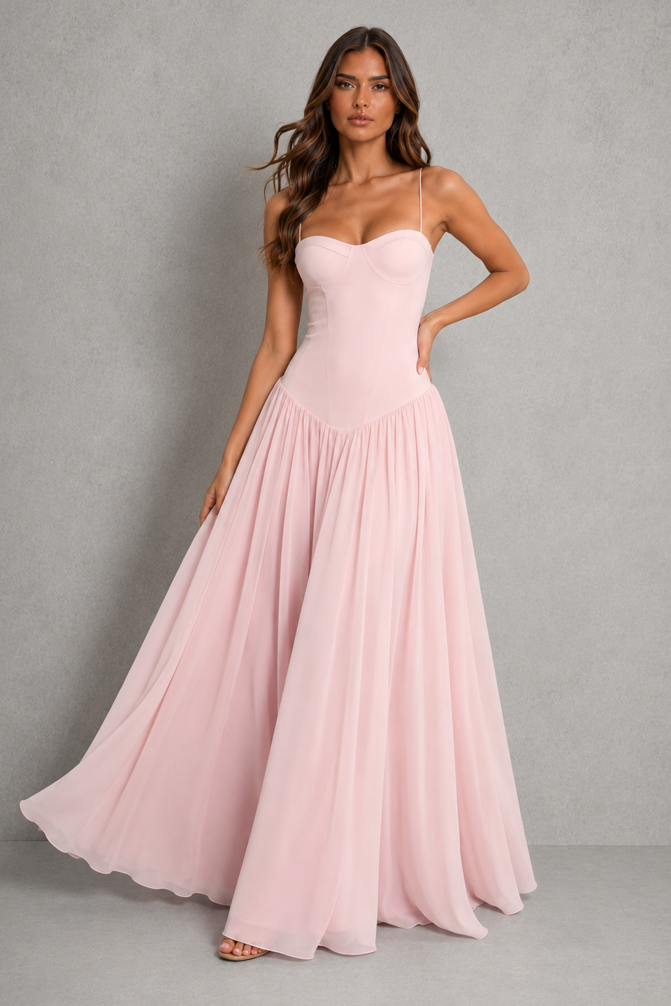 A woman modeling a light pink strapless maxi dress with a fitted bodice and a flowing chiffon skirt, standing against a textured gray studio background.