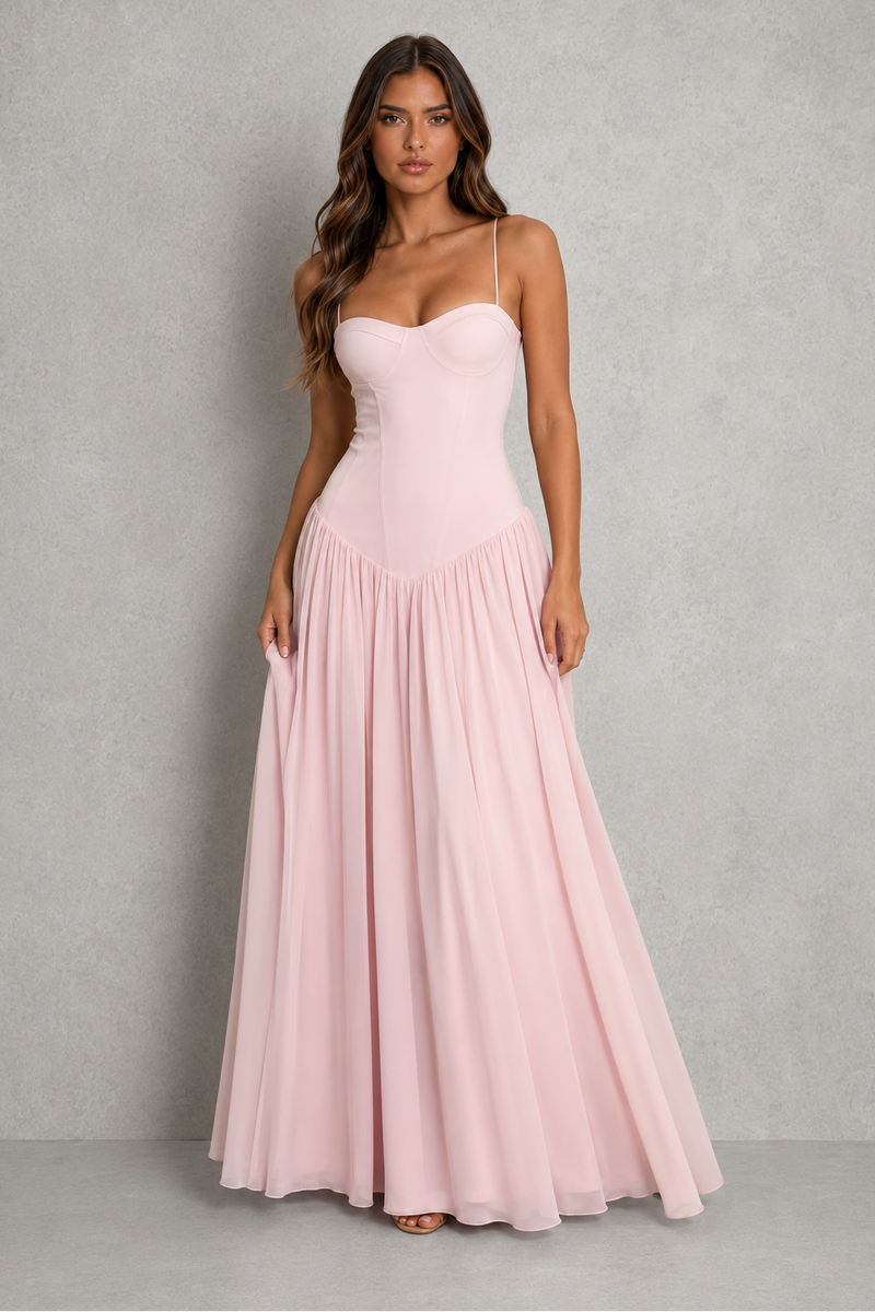 A woman modeling a light pink sleeveless maxi dress with a sweetheart neckline, fitted bodice, and flowing chiffon skirt, standing against a textured gray studio backdrop.