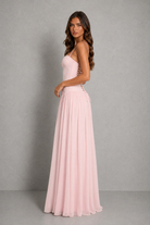 A woman modeling a floor-length light pink spaghetti-strap dress with a fitted bodice and flowing skirt, standing against a neutral gray studio backdrop.