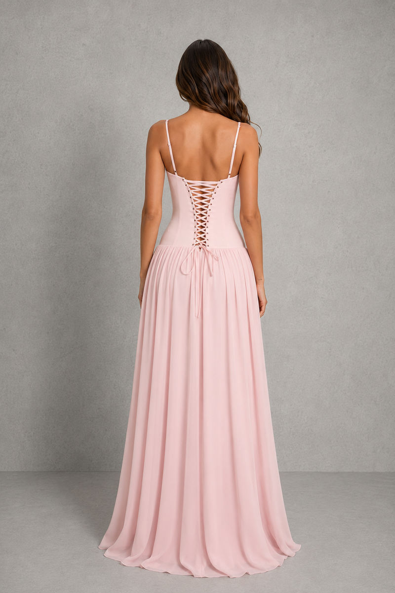 A woman viewed from the back wearing a long blush pink dress with thin straps, a lace-up open back, and a flowing skirt, standing against a plain gray studio backdrop.