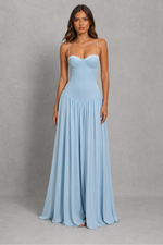 A woman modeling a light blue sleeveless maxi dress with a sweetheart neckline, thin spaghetti straps, a structured corset-style bodice, and a flowing pleated skirt, standing against a textured gray studio backdrop.