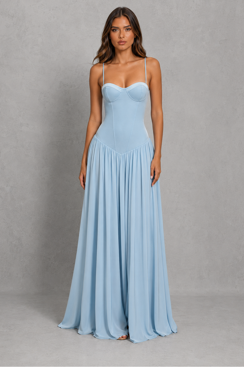 A woman modeling a light blue sleeveless maxi dress with a sweetheart neckline, thin spaghetti straps, a structured corset-style bodice, and a flowing pleated skirt, standing against a textured gray studio backdrop.