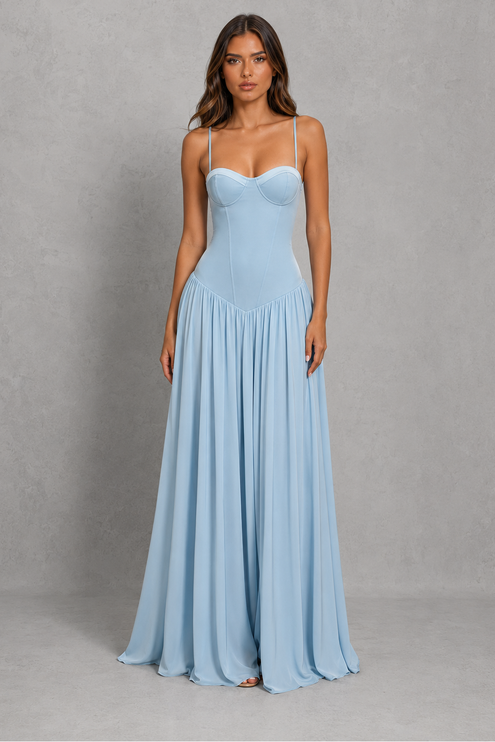 A woman modeling a light blue sleeveless maxi dress with a sweetheart neckline, thin spaghetti straps, a structured corset-style bodice, and a flowing pleated skirt, standing against a textured gray studio backdrop.