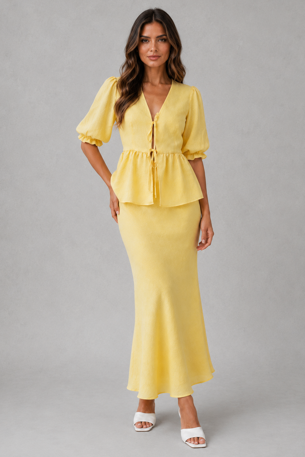 A woman modeling a two-piece yellow dress set consisting of a short-sleeve peplum top with a tie-front detail and a matching high-waisted mermaid-style skirt, paired with white heeled sandals against a neutral gray studio background.