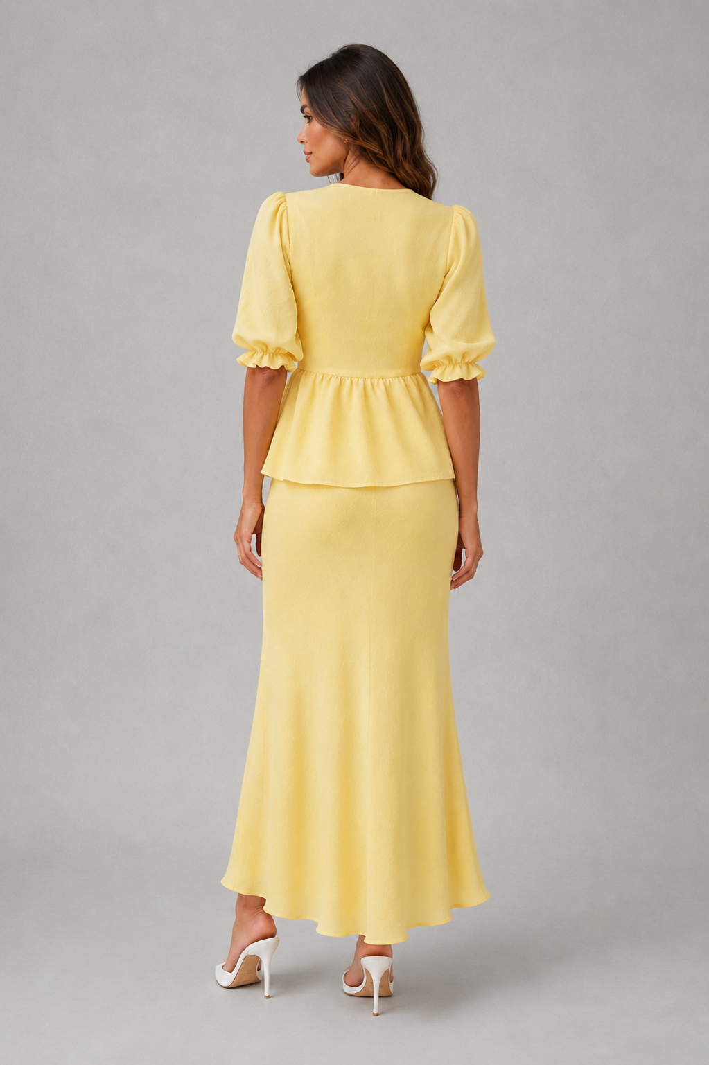 A woman shown from the back wearing a pale yellow peplum maxi dress with short puff sleeves and white high-heeled sandals, photographed against a plain light gray studio background.