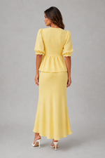 A woman shown from the back wearing a pale yellow peplum maxi dress with short puff sleeves and white high-heeled sandals, photographed against a plain light gray studio background.