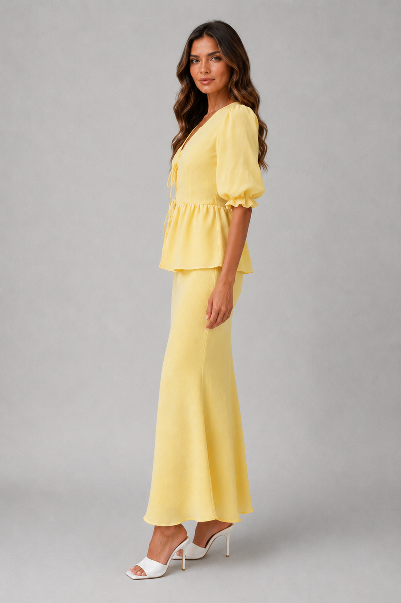 A woman modeling a two-piece yellow outfit consisting of a short puff-sleeve peplum top and a matching long skirt, paired with white heeled sandals, against a solid gray studio background.