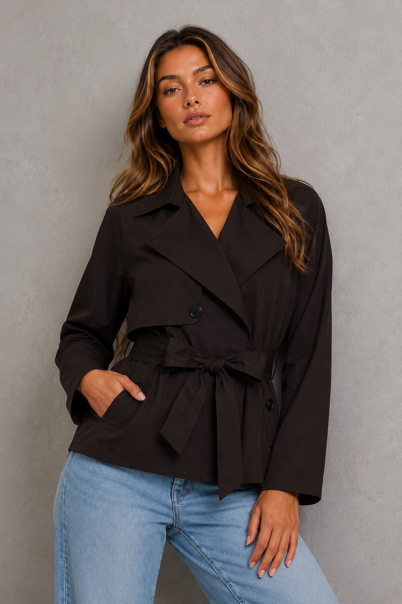 A woman modeling a dark brown belted wrap jacket with a collar and front buttons, paired with light blue jeans, standing against a textured gray wall.