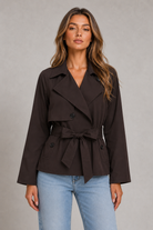 A woman modeling a dark brown double-breasted belted jacket with light wash blue jeans, standing against a neutral gray studio background.