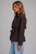 A woman wearing a dark brown belted wrap jacket and light blue jeans, shown in profile against a plain gray studio background.