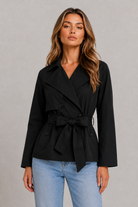 A woman modeling a black belted wrap jacket with a collar and front buttons, paired with light blue jeans, against a neutral gray studio background.
