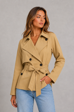 A woman modeling a tan double-breasted belted trench jacket with black buttons, paired with light blue jeans, against a neutral gray studio background.