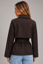 A woman shown from the back wearing a dark brown cropped jacket with a matching waist belt, paired with light blue jeans, against a solid gray studio background.