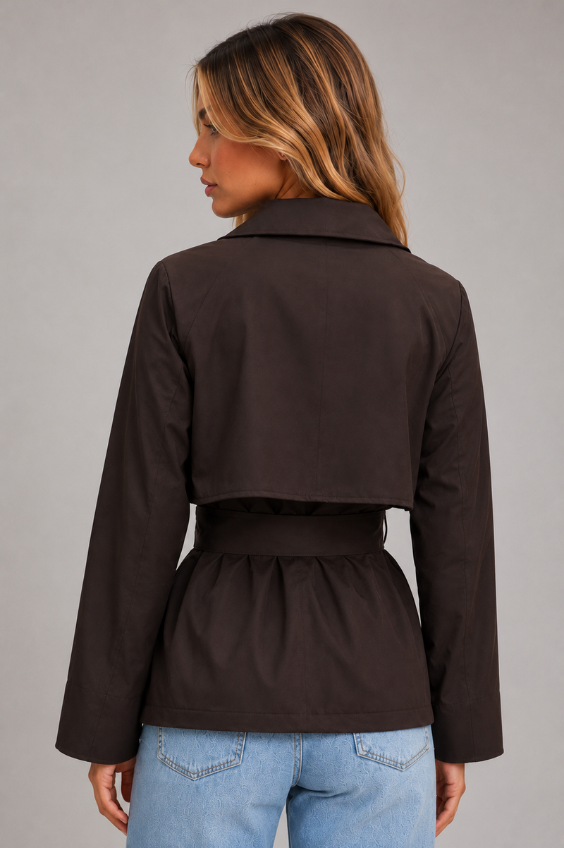 A woman shown from the back wearing a dark brown cropped jacket with a matching waist belt, paired with light blue jeans, against a solid gray studio background.