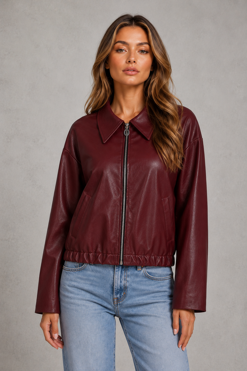 A woman modeling a cropped, long-sleeve burgundy faux-leather jacket with a front zipper and pointed collar, paired with light-wash blue jeans against a textured gray studio background.