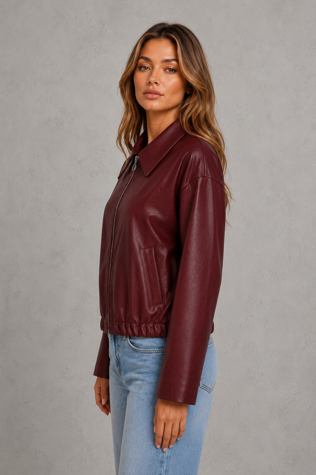 A woman wearing a burgundy faux-leather jacket and light blue jeans, standing against a textured gray studio background.