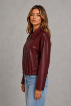 A woman wearing a burgundy faux-leather jacket and light blue jeans, standing against a textured gray studio background.