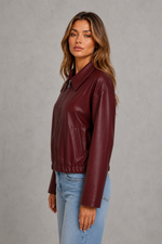 A woman wearing a burgundy faux-leather jacket and light blue jeans, standing against a textured gray studio background.