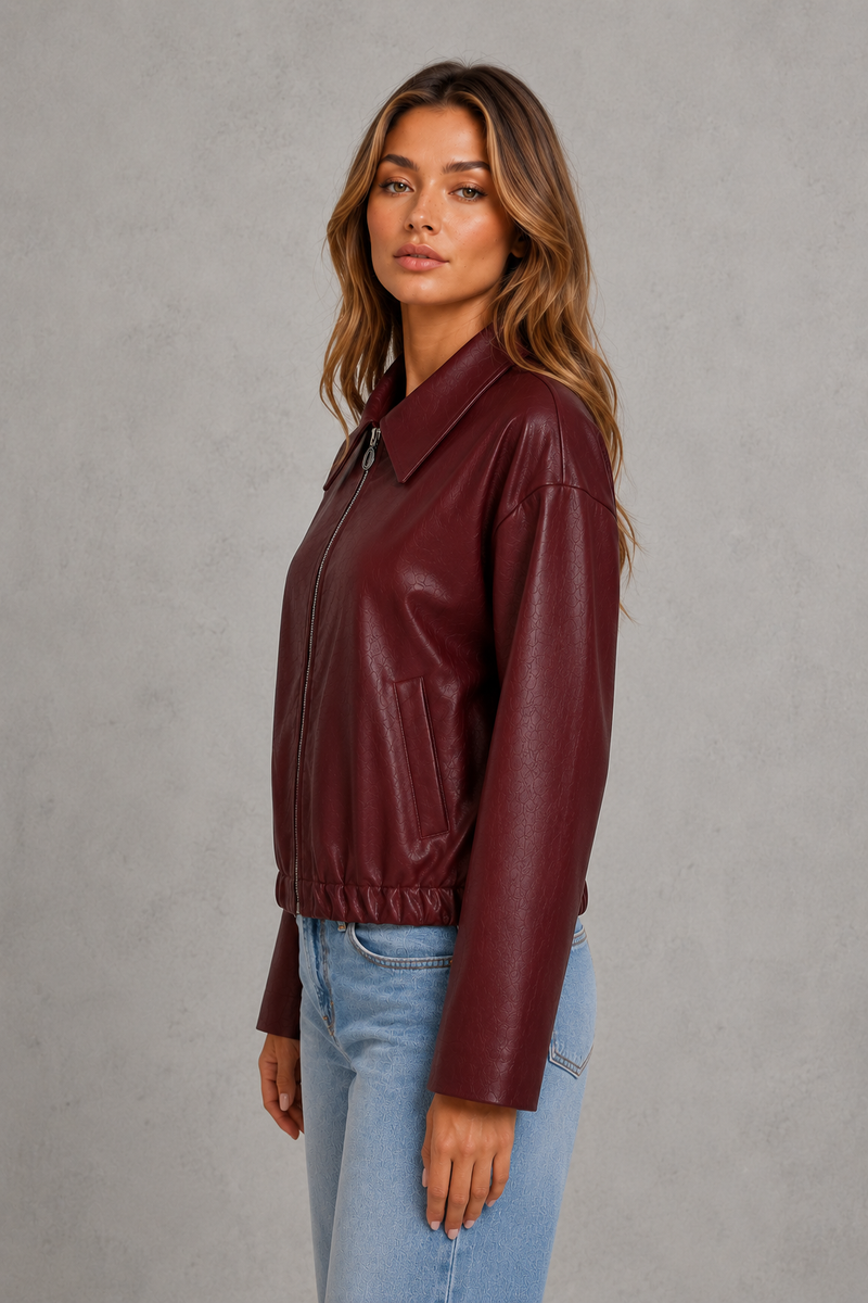 A woman wearing a burgundy faux-leather jacket and light blue jeans, standing against a textured gray studio background.