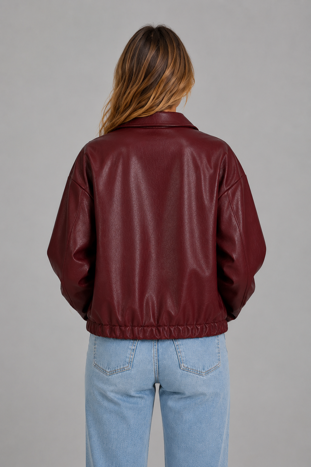 A woman viewed from behind wearing a cropped, long-sleeve burgundy faux-leather jacket and light blue jeans against a solid gray studio background.