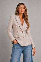 A woman wearing a light beige textured double-breasted blazer with gold buttons and blue jeans, standing against a gray concrete wall.