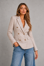 A woman wearing a light beige textured double-breasted blazer with gold buttons and blue jeans, standing against a gray concrete wall.