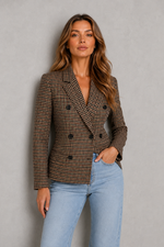 A woman modeling a brown and beige houndstooth double-breasted blazer with light blue jeans, standing against a textured gray wall.