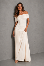 An off-the-shoulder, floor-length pleated maxi dress in cream or off-white, modeled against a textured gray studio backdrop.