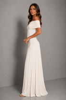 A woman modeling an off-the-shoulder, floor-length pleated dress in a warm off-white or cream color, standing against a neutral gray studio backdrop.