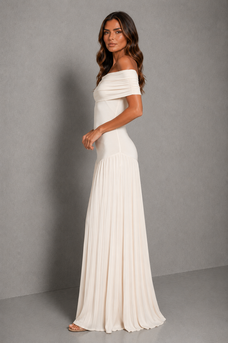 A woman modeling an off-the-shoulder, floor-length pleated dress in a warm off-white or cream color, standing against a neutral gray studio backdrop.