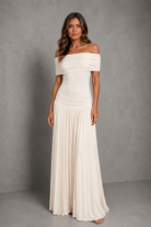 An off-the-shoulder floor-length gown in a warm ivory or cream color, featuring a ruched bodice and a flowing pleated skirt, modeled against a textured gray studio backdrop.