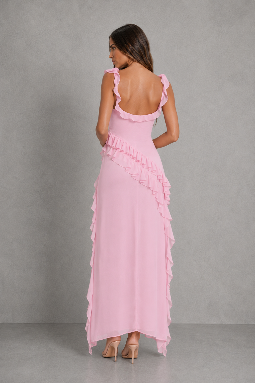 A woman modeling a light pink maxi dress from the back, featuring ruffled shoulder straps, a ruffled waistline, and cascading ruffle details down the sides, set against a textured gray studio background.