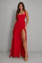 A woman modeling a bright red one-shoulder maxi dress with ruffle details and a high leg slit, standing against a textured gray studio backdrop.