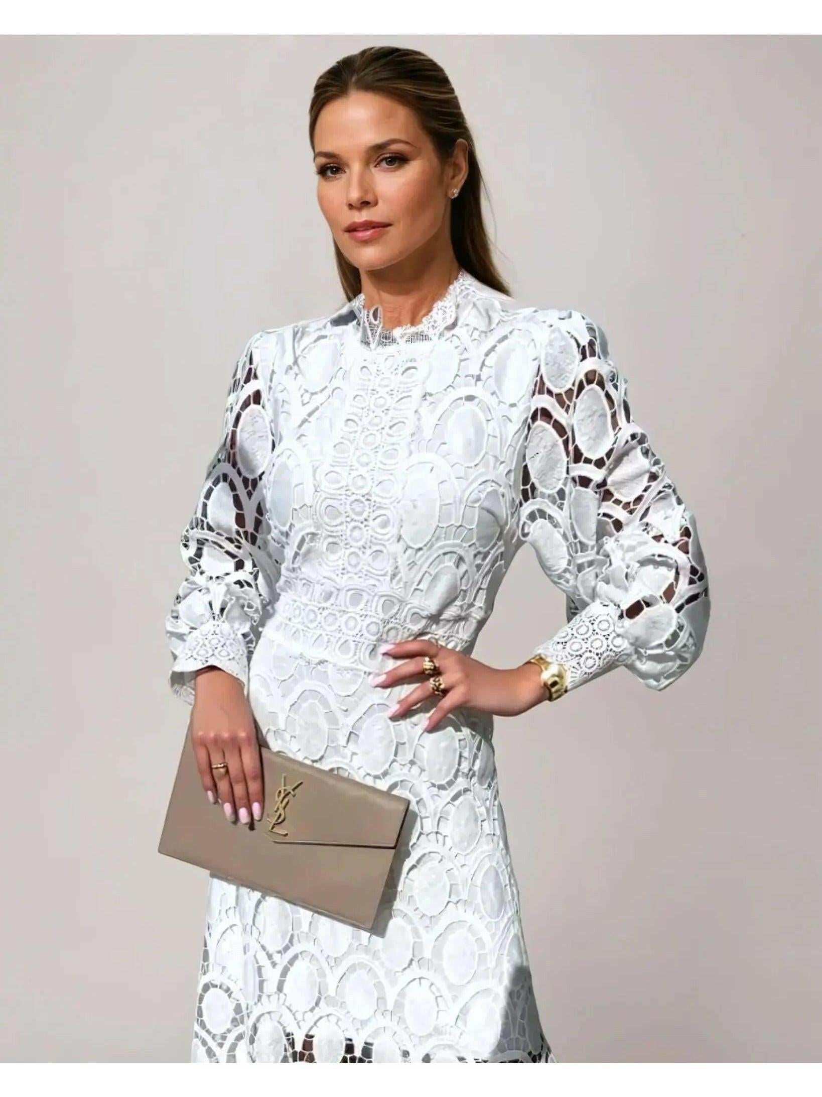 Woman wearing a white lace dress holding a beige clutch against a plain background