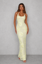 A woman modeling a pale yellow floral lace halter-neck maxi dress against a textured gray studio backdrop.