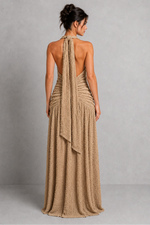 Woman wearing a beige halter neck dress with a draped back on a plain background